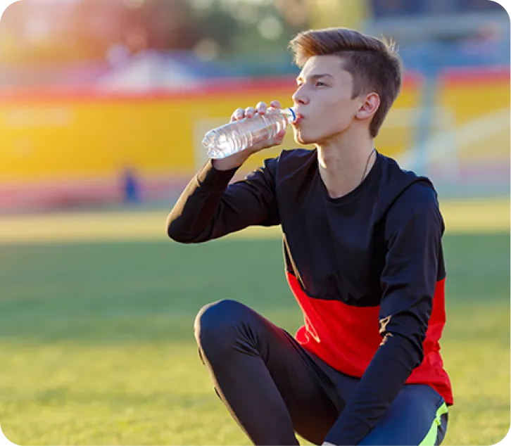 Accroupi sur un terrain de sport, un adolescent boit dans une bouteille d’eau jetable.